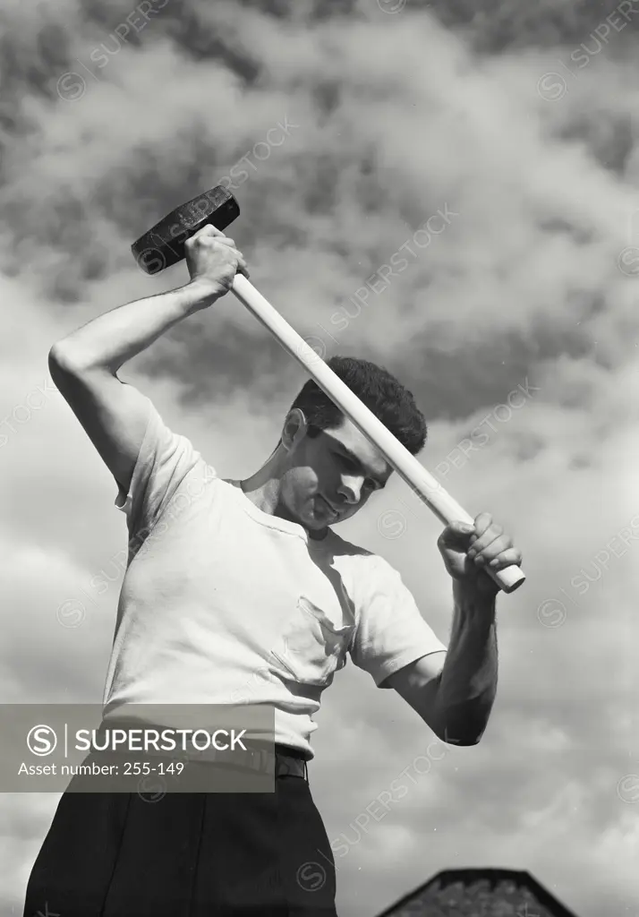 Vintage Photograph. Looking up at man wearing white t-shirt swinging sledgehammer outside with clouds in sky