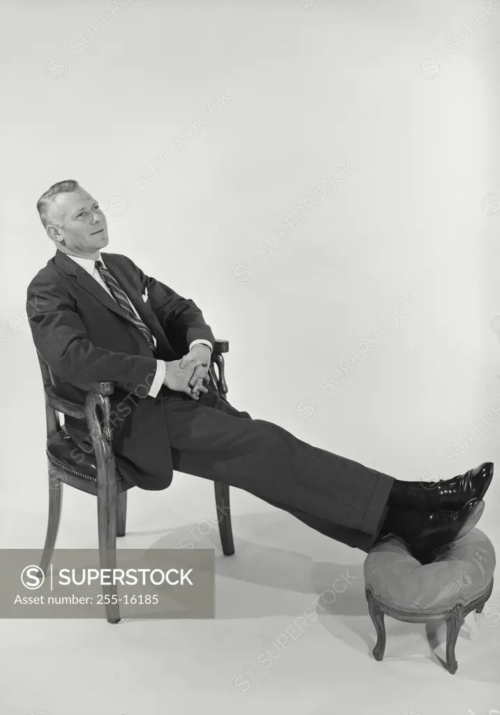 Vintage Photograph. Man wearing suit sitting in chair with feet perched on footstool on white background, Frame 2