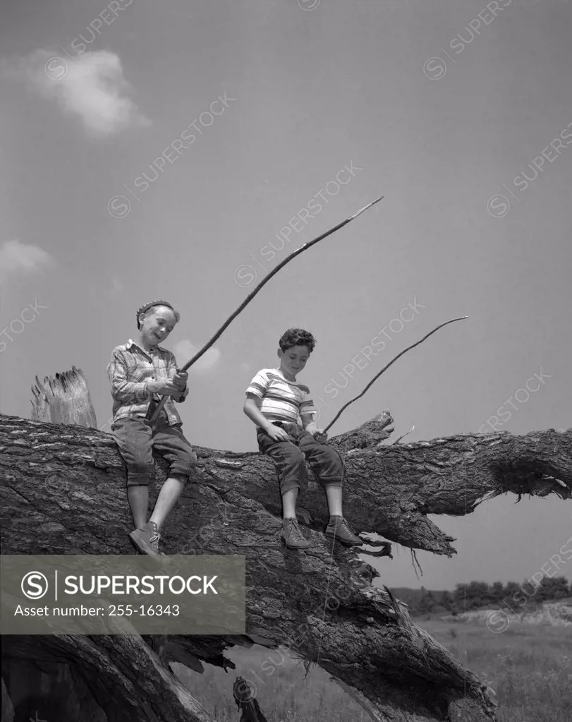 Two boys sitting on tree trunk and fishing with homemade fishing rod