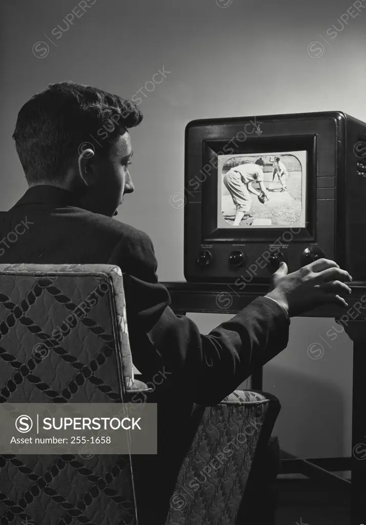Vintage Photograph. Young man sitting in chair watching vintage television set showing baseball players on screen