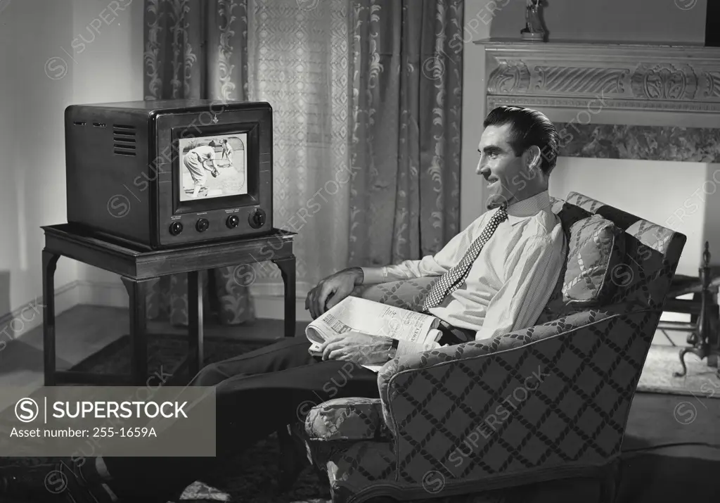 Vintage Photograph. Man relaxing in chair in living room with newspaper in lap watching television with baseball players on screen
