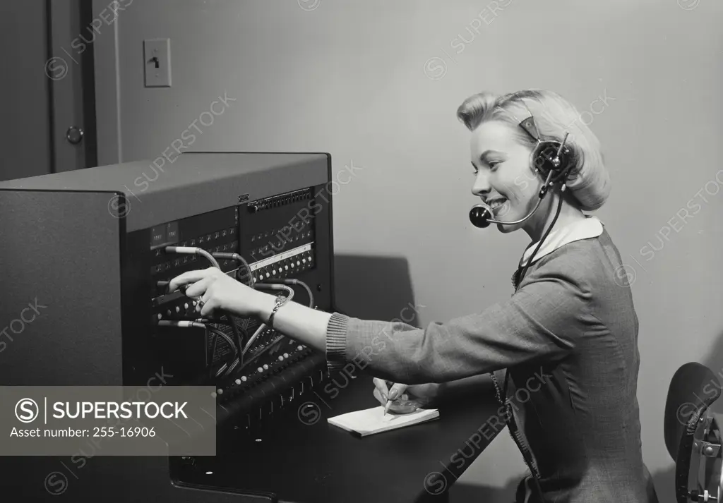 Vintage Photograph. Blonde woman switchboard operator smiling wearing headset and writing in notepad, Frame 1