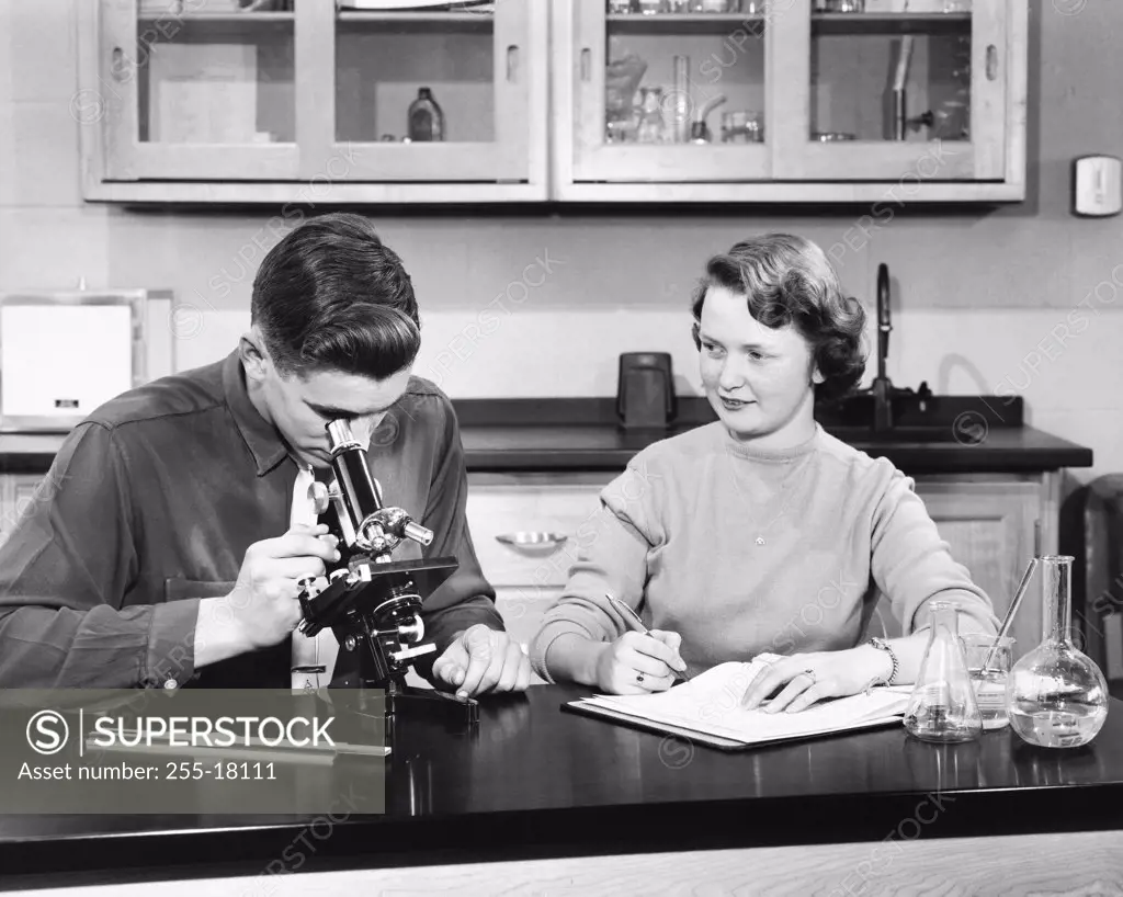 Teenage boy looking through a microscope with a teenage girl sitting beside him in a laboratory