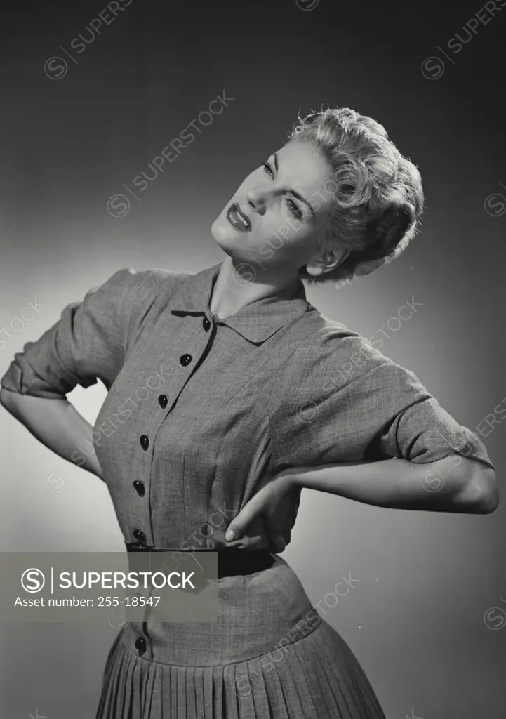 Vintage Photograph. Blonde woman with short hair wearing dress with black buttons and black belt holding hands on hips looking up in thought