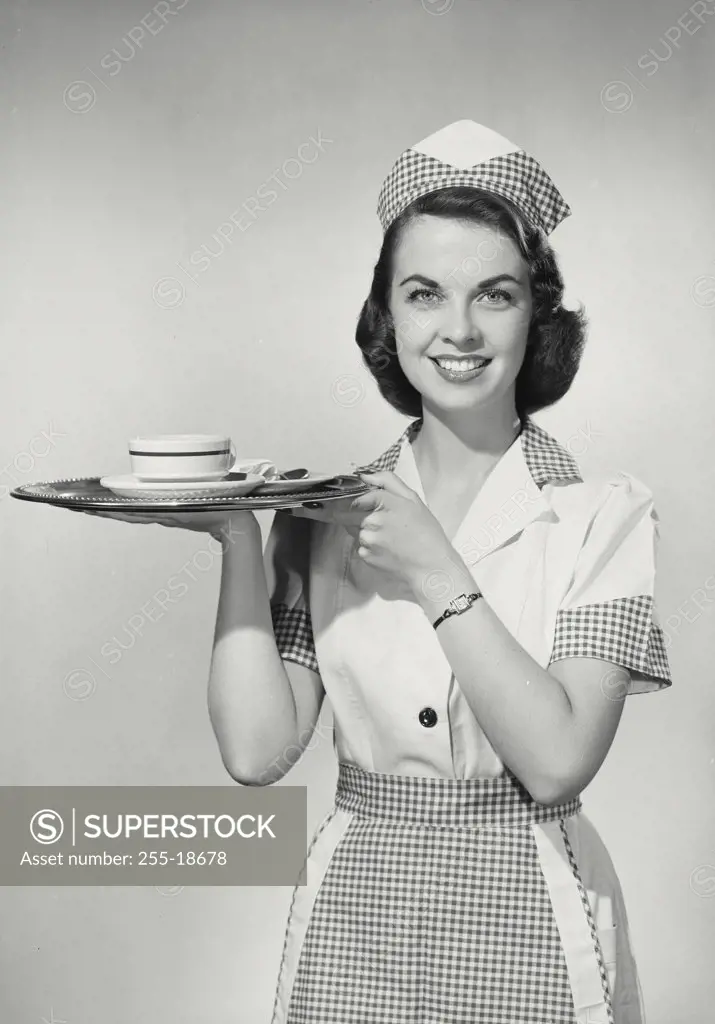 Vintage photograph. Brunette woman wearing waitress uniform holding tray with coffee cup to shoulder turned to camera and smiling