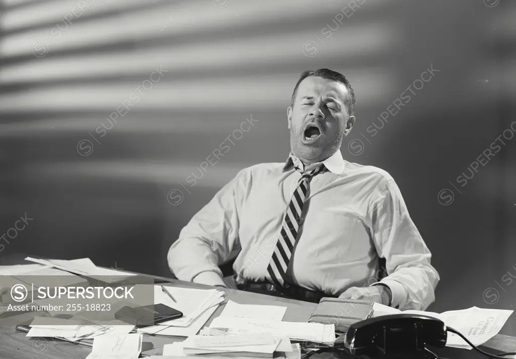 Vintage Photograph. Man in dress shirt and striped tie sitting at desk covered with papers yawning