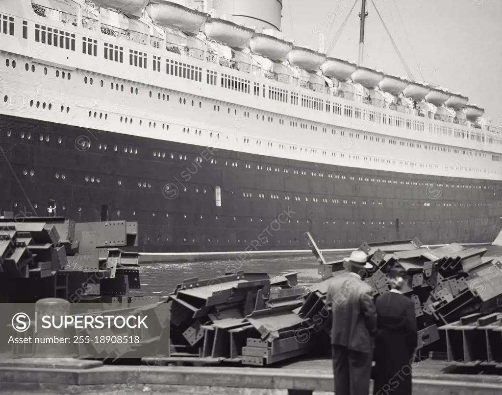 Vintage photograph. Couple in front of steel beams on dock with Queen Elizabeth ship in background