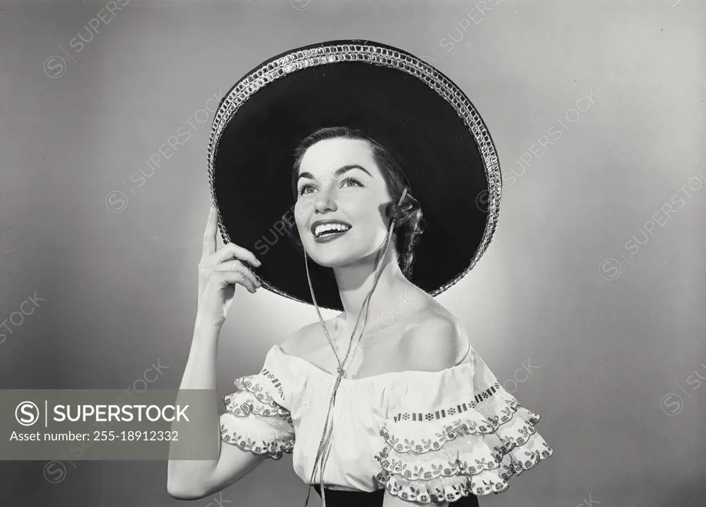Vintage photograph. Brunette woman wearing traditional Mexican dress and hand raised to black sombrero with silver trim detail