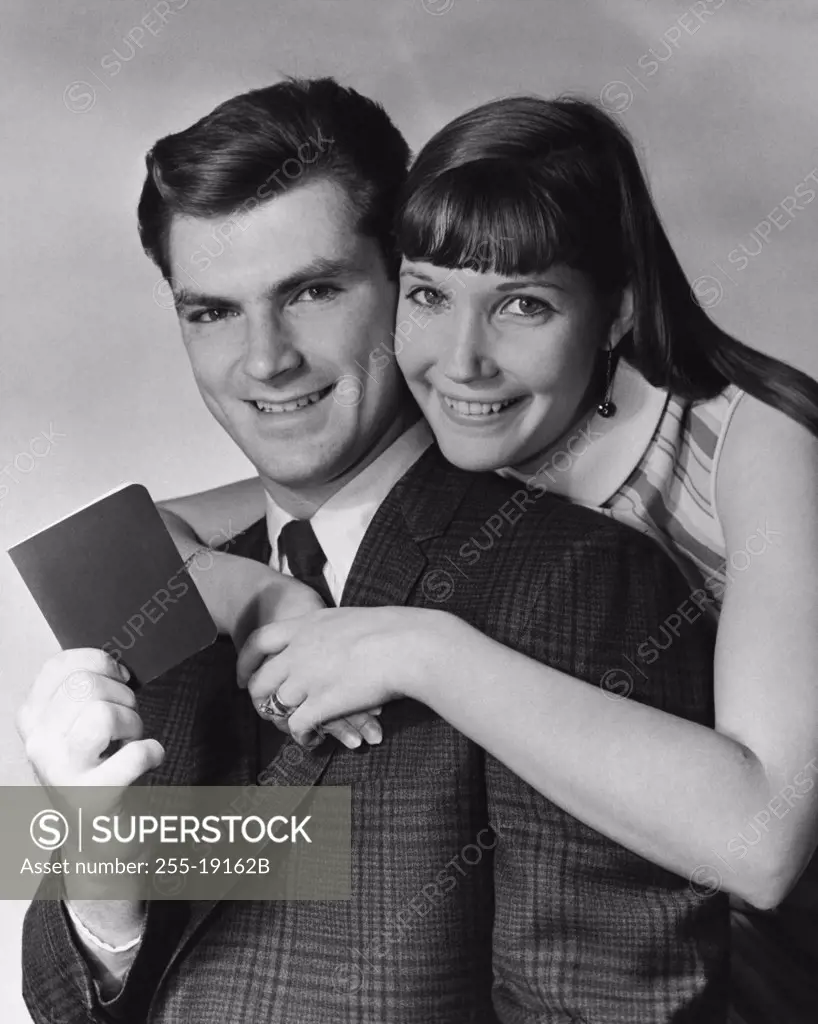 Portrait of a young man holding a bankbook with a young woman embracing him from behind