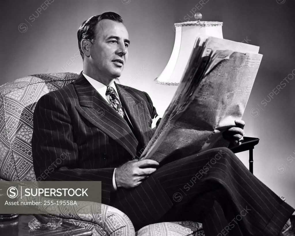 Vintage Photograph. Dark haired caucasian man wearing pin striped suit sitting in chair reading newspaper