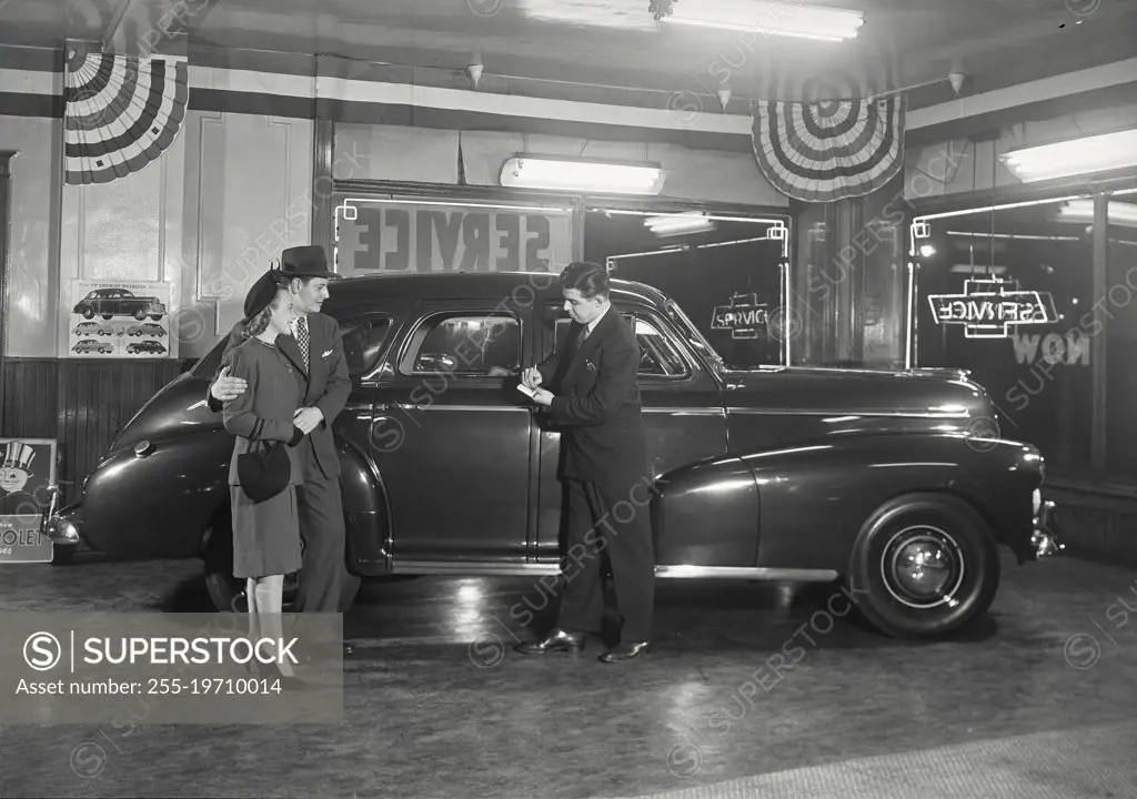 Vintage photograph. Couple in car dealership standing next to Chevrolet Stylemaster talking to salesman