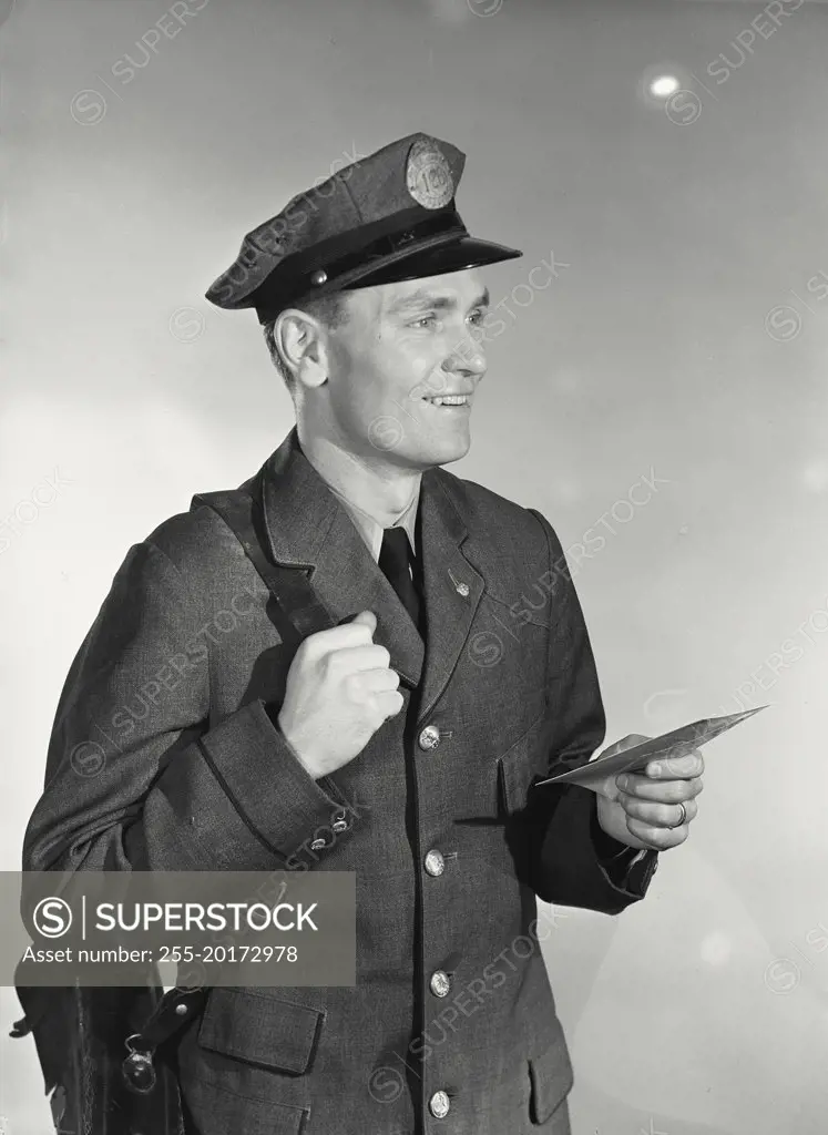 Vintage photograph. Mailman wearing uniform, hat and satchel bag holding up letter looking to side