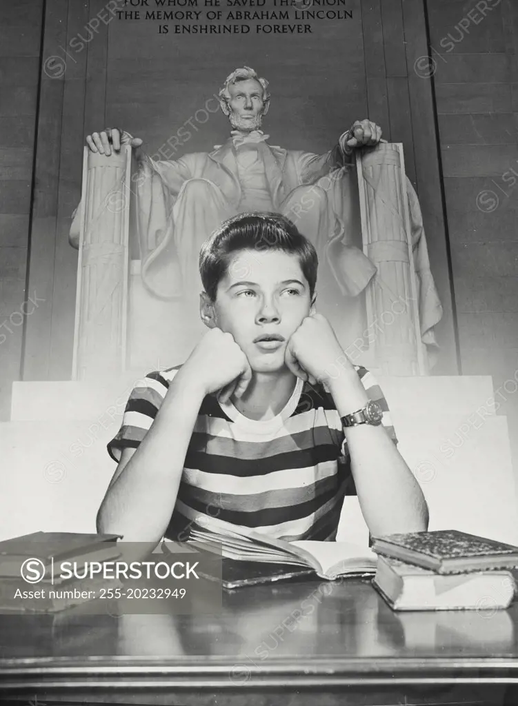 Boy at desk with book open resting cheek on fist with the Lincoln Memorial behind 