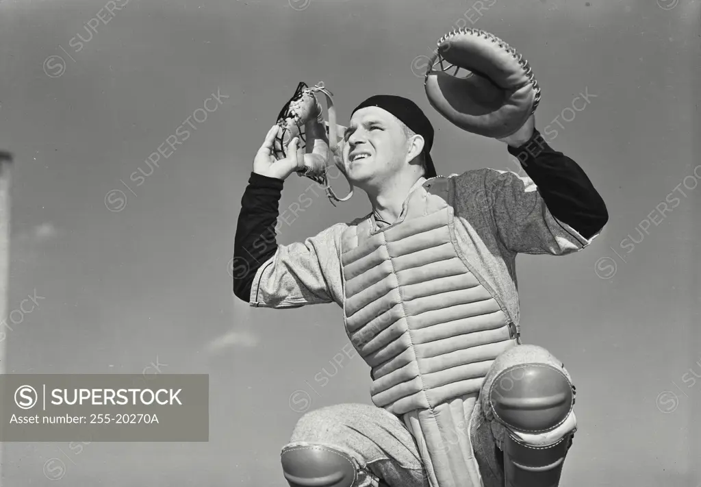 Vintage Photograph. Man wearing baseball catcher uniform taking mask off looking out holding up catcher's mitt