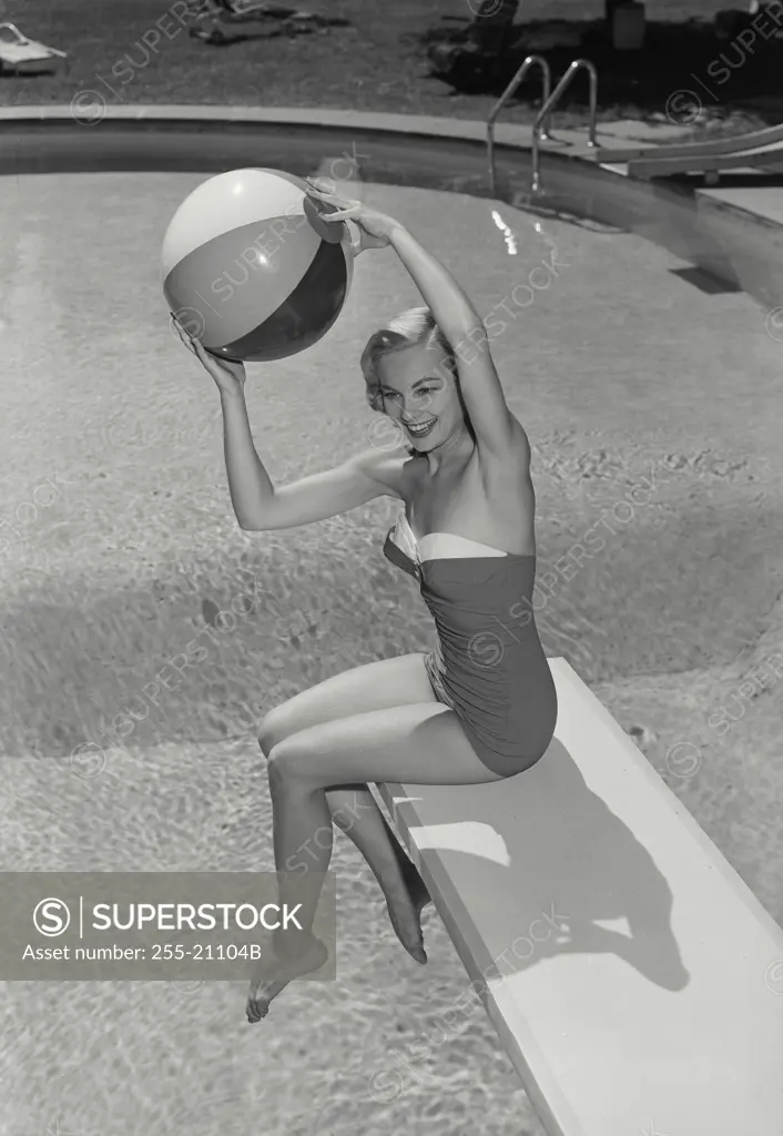 Vintage Photograph. Smiling blonde woman sitting on diving board near swimming pool holding up beach ball over head