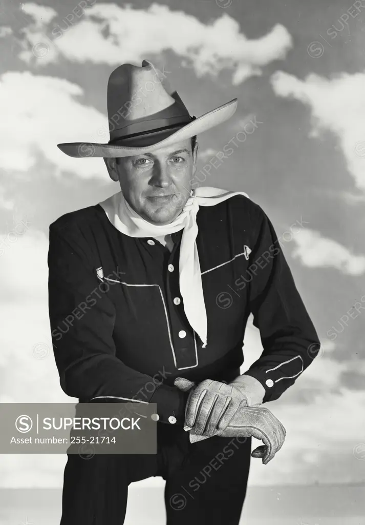 Vintage Photograph. Man wearing black cowboy outfit with hat and white bandana kneeling in front of cloud sky background