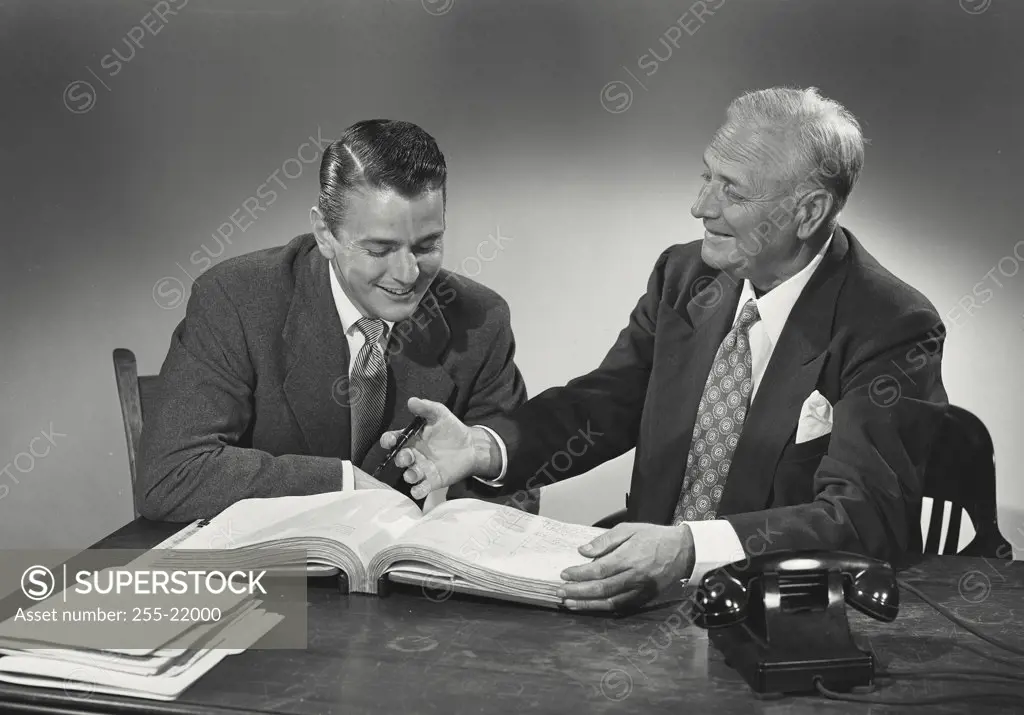 Young man and older man sitting together at desk reading through book