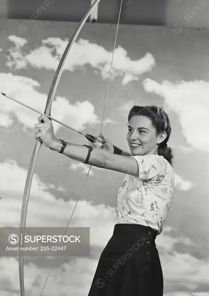 Vintage Photograph. Smiling young woman turned to side aiming bow and arrow in front of cloud sky background
