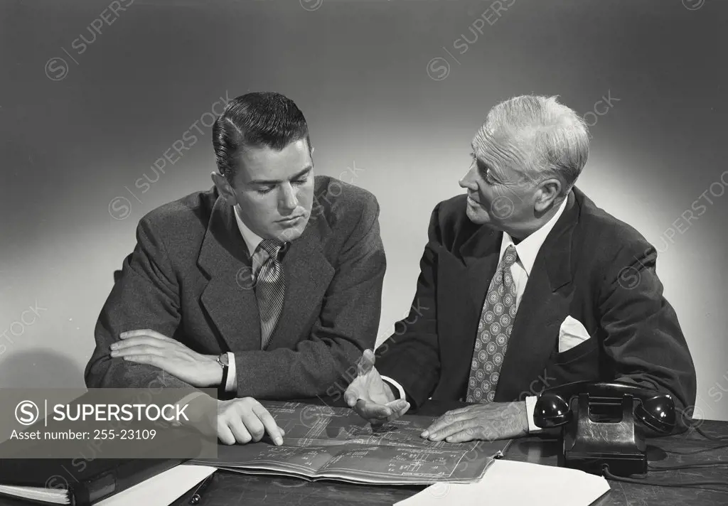 Young man and older man sitting together at desk looking over blueprints