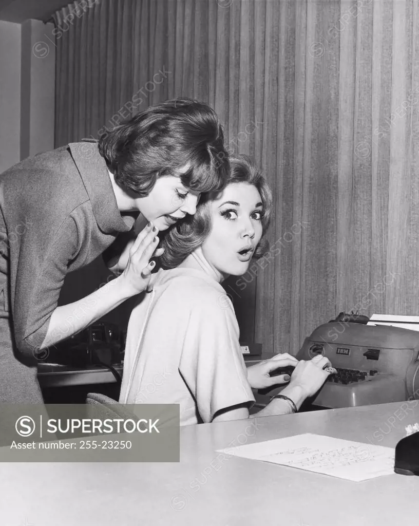 Side profile of a businesswoman whispering to another businesswoman in an office