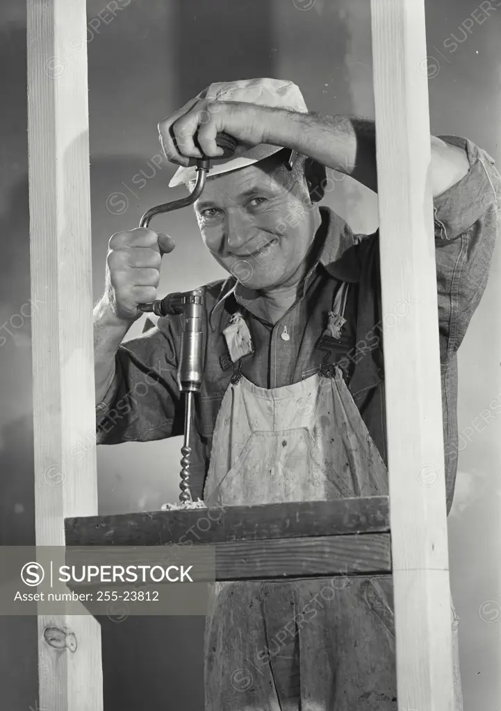 Vintage Photograph. Older carpenter using hand powered drill on blocking board between wooden studs smiling at camera