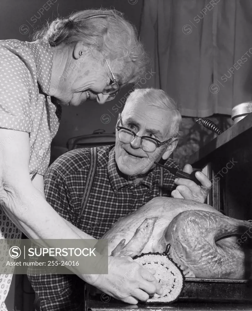 Side profile of a senior woman putting a turkey into an oven with a senior man looking at her