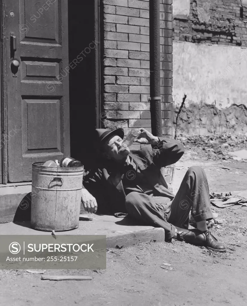 High angle view of a homeless senior man drinking alcohol sitting near a garbage can