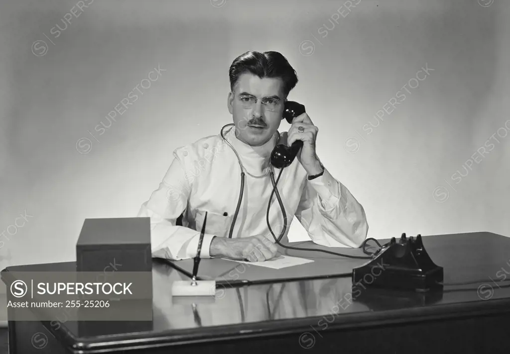 Vintage Photograph. Male doctor with dark hair and mustache and stethoscope around neck sitting at desk holding telephone reciever to ear