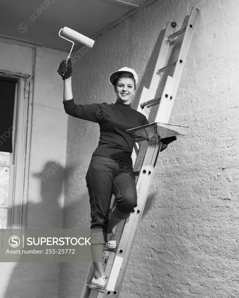 Portrait of a young woman standing on a ladder and holding a paint roller and a paint tray