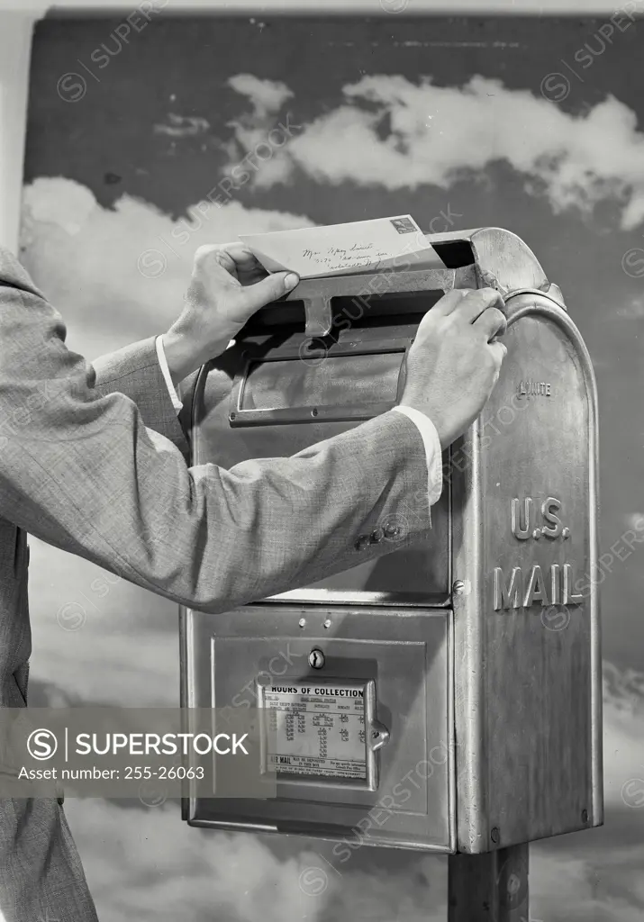Vintage Photograph. Man wearing suit placing envelopes into slot of US Mail box with cloud sky background
