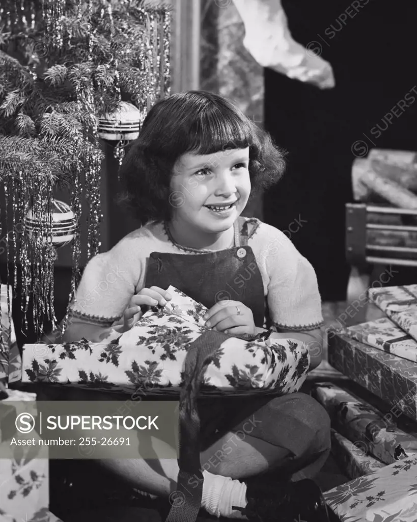Vintage Photograph. Young girl opening Christmas gift next to tree and smiling