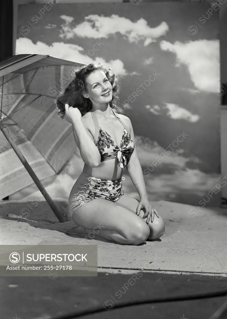 Vintage Photograph. Smiling curly haired woman wearing swimsuit sitting in sand on beach set with large umbrella and sky backdrop playing with hair
