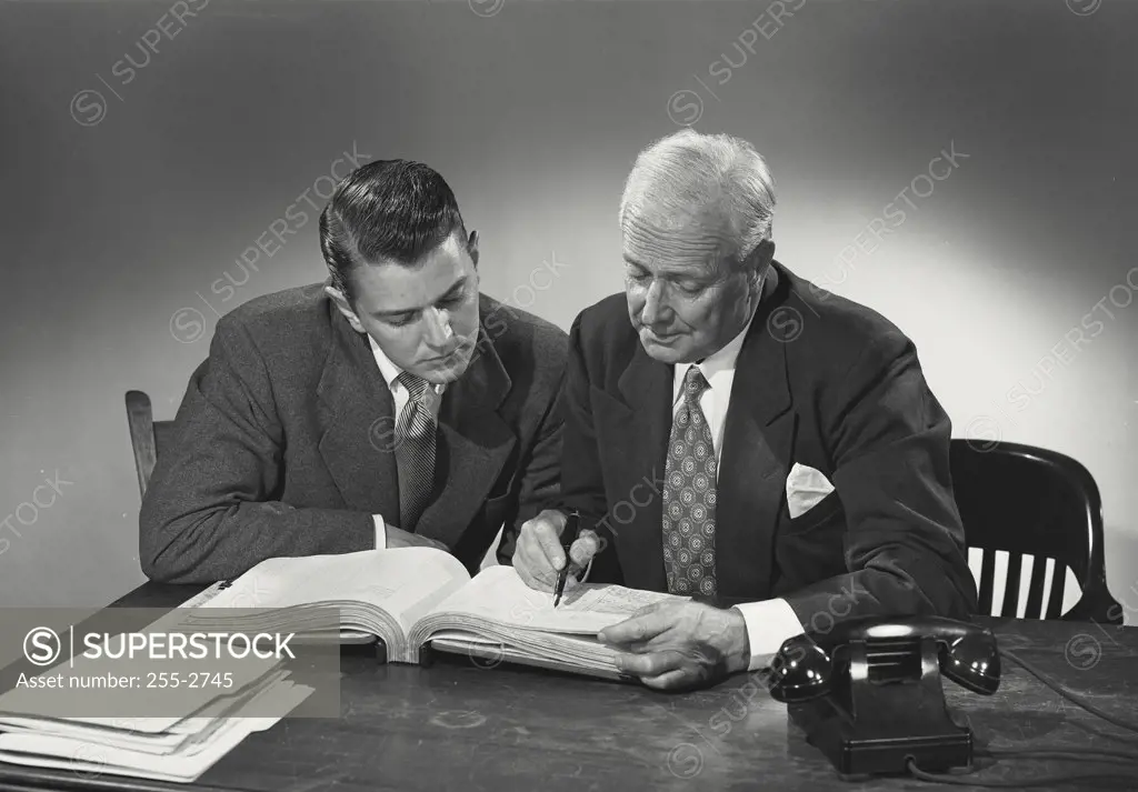 Young man and older man sitting together at desk reading through book