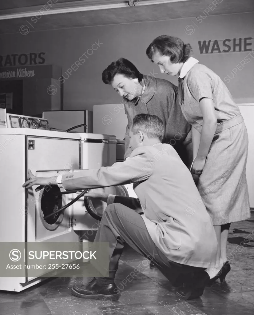 Side profile of a salesman showing a washing machine to two female customers