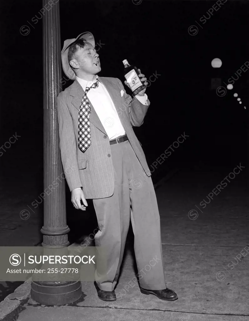 Drunk mid adult man leaning against a lamppost holding a bottle of alcohol