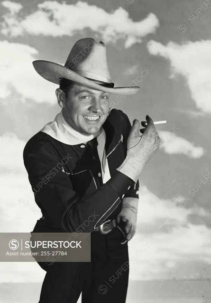Vintage Photograph. Smiling man wearing black cowboy outfit with hat and white bandana kneeling in front of cloud sky background with cigarette