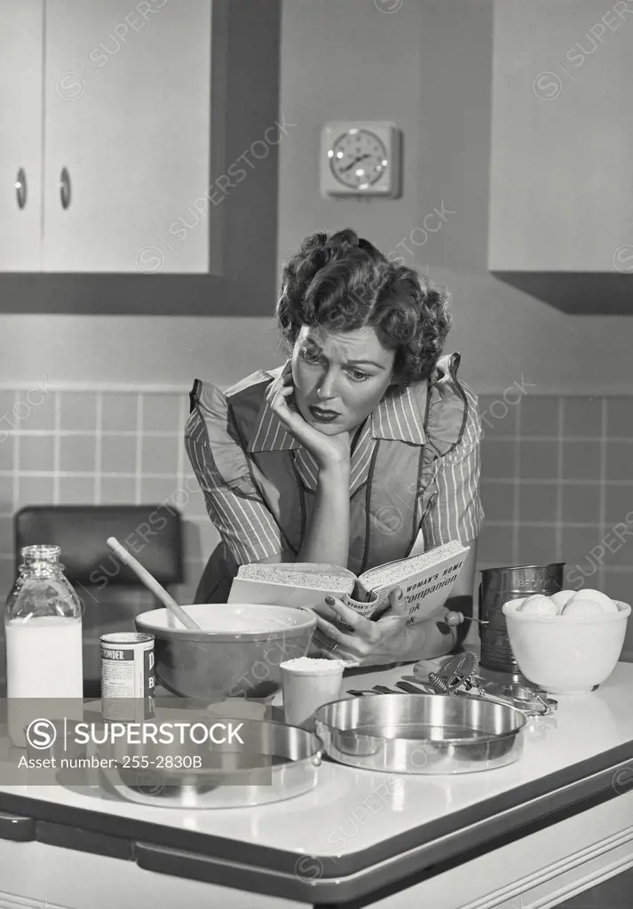 Vintage photograph. Mid adult woman reading a cookbook in a domestic kitchen