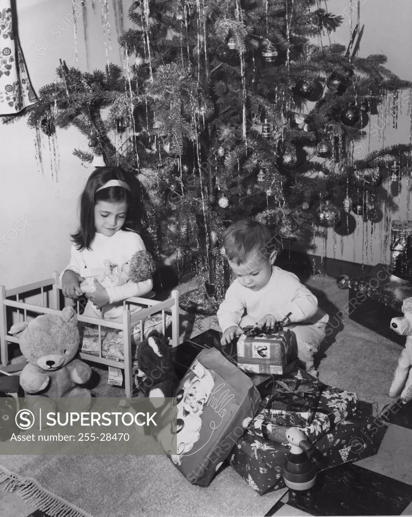 Girl playing with a doll and her brother opening a Christmas present beside her, 1960s