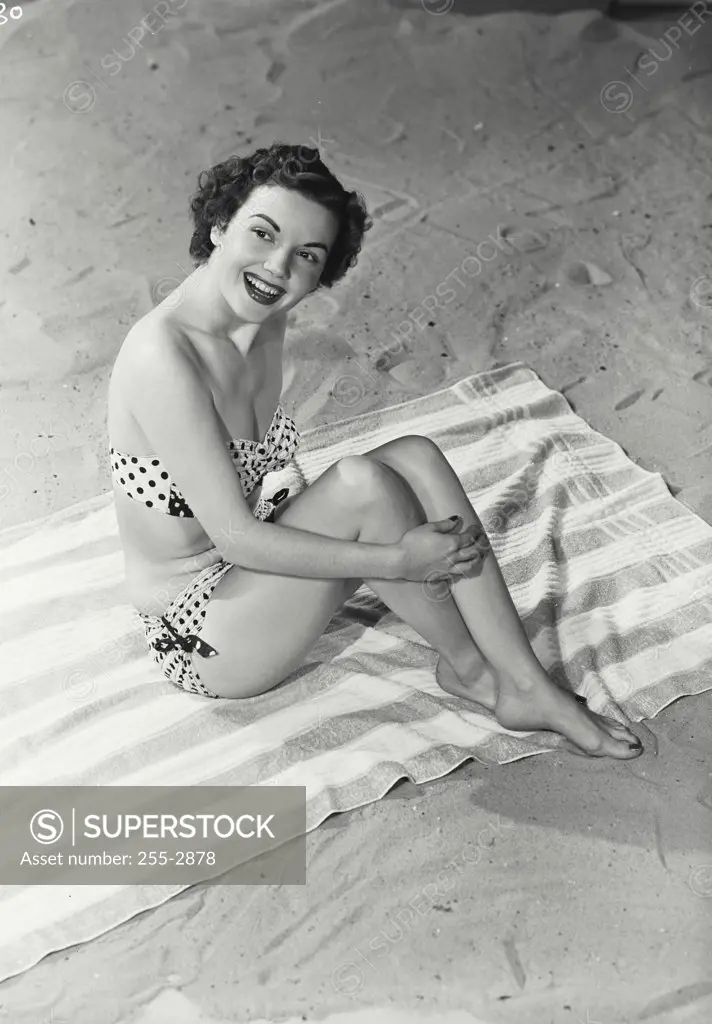 Vintage photograph. High angle view of a young woman smiling in swimsuit sitting on towel in sand.