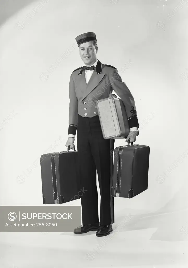 Vintage Photograph. Smiling man wearing bellman uniform standing on white background holding three suitcases