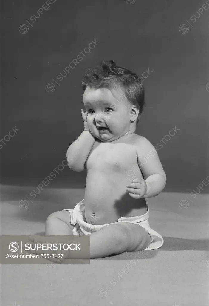 Vintage Photograph. Baby girl sitting on blanket on flat surface holding up arm near cheek