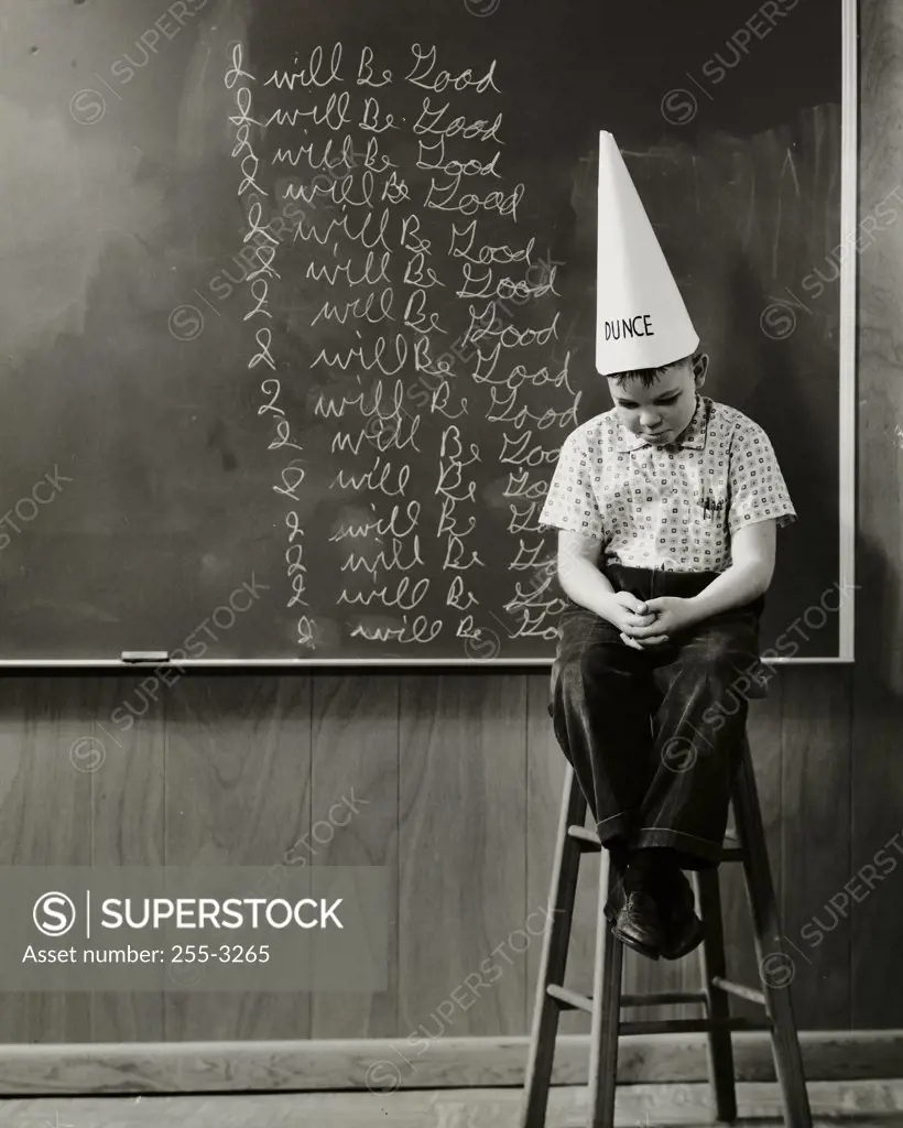 Boy wearing a dunce cap sitting in front of a blackboard