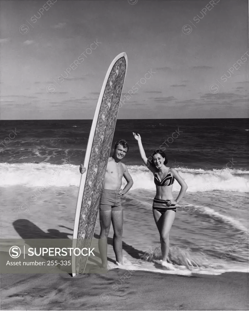 Young man holding surfboard on beach with young woman standing beside him