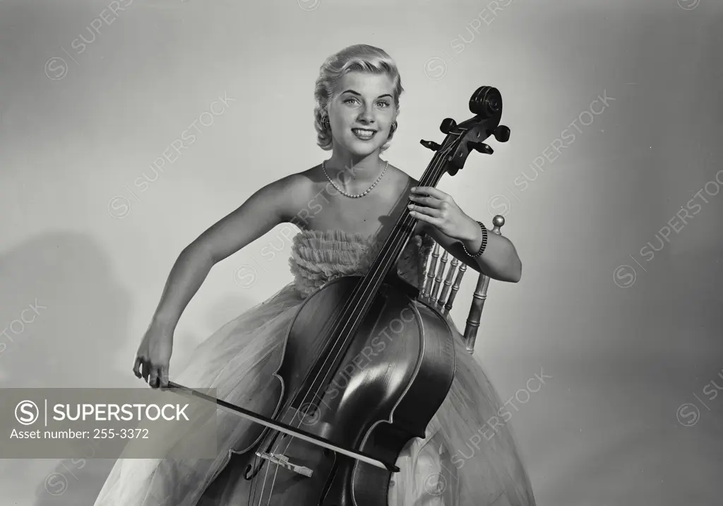 Vintage Photograph. Young blonde woman wearing evening gown sitting in wooden chair holding cello and bow in front of solid studio backdrop, Frame 5