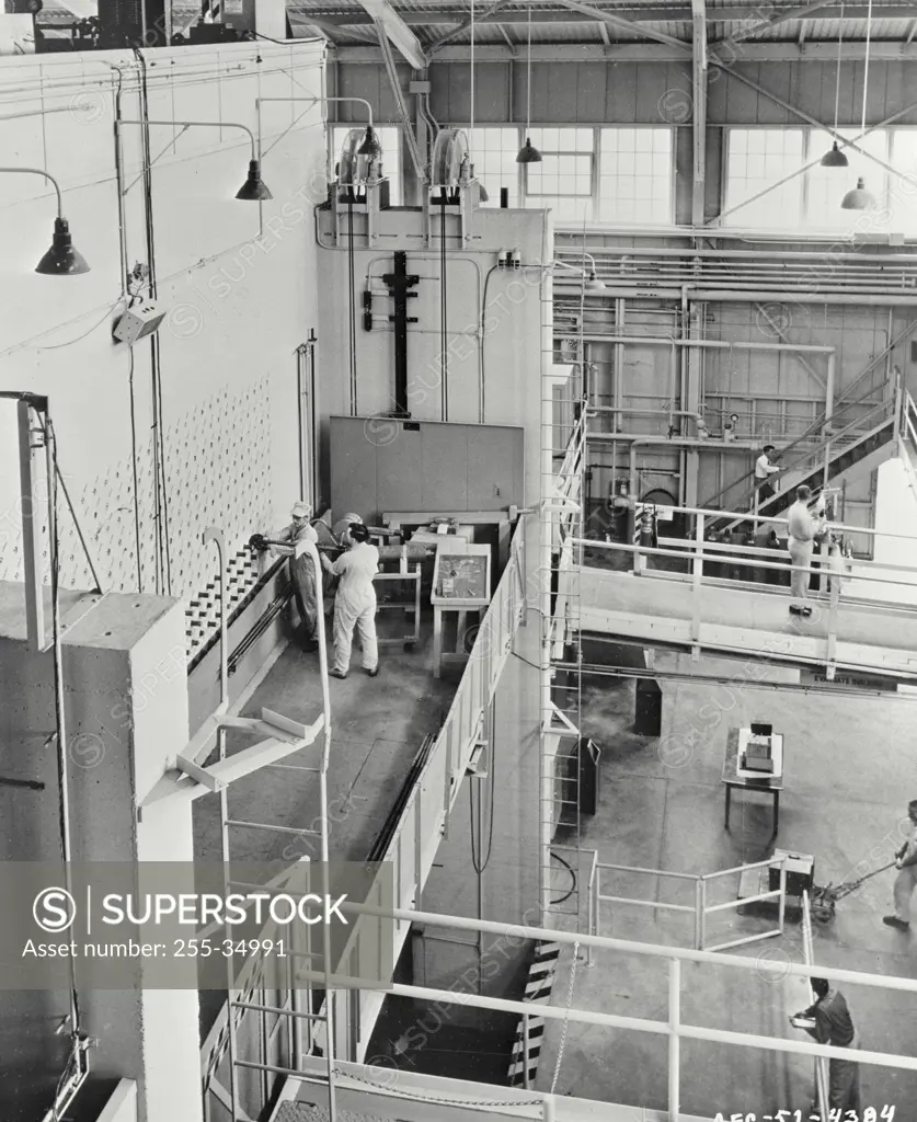 Vintage photograph. Preparing to remove plugs from some of the 1,248 fuel channel openings in the shield of the Oak Ridge graphite reactor, personnel stand on elevating platform. In front of the two men at top left, can be seen a horizontal bundle of 10 foot lengths of light steel poles used as push rods