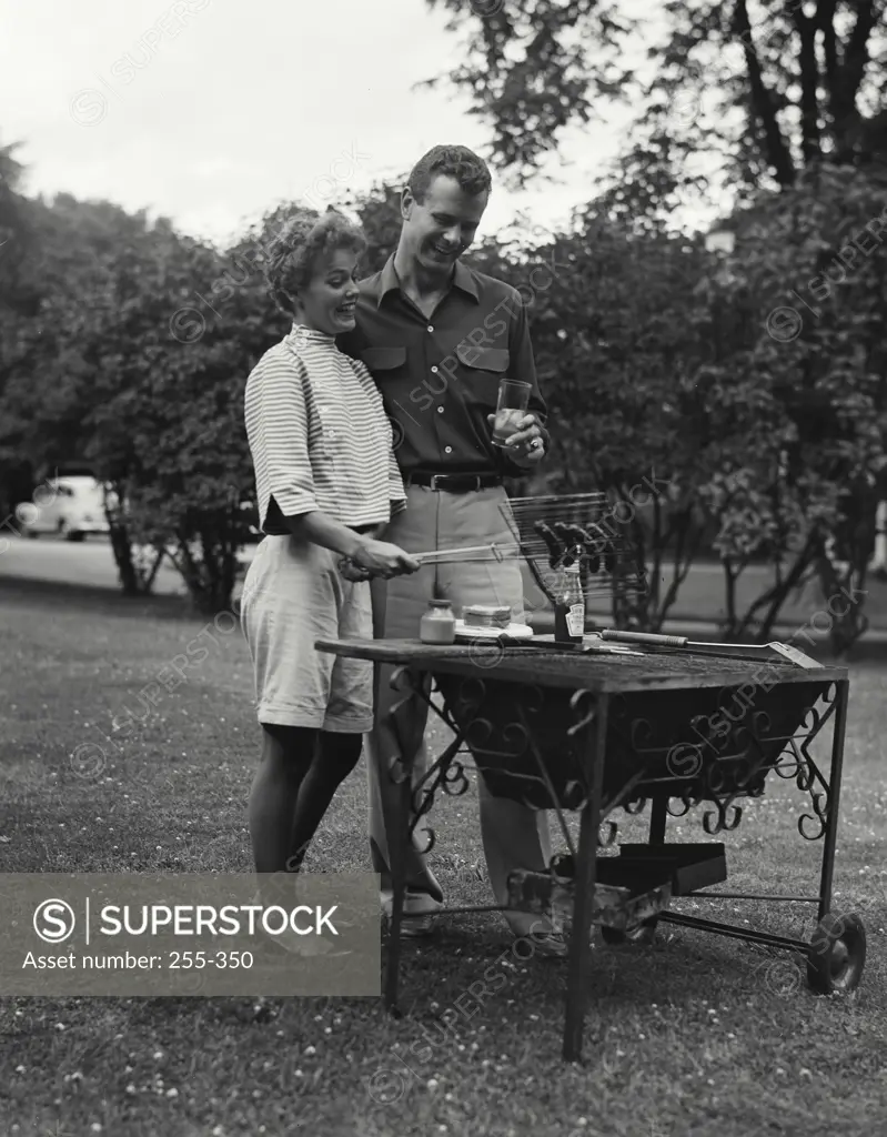 Vintage Photograph. Young couple - woman outside at park picnic grill roasting frankfurters as man looks on