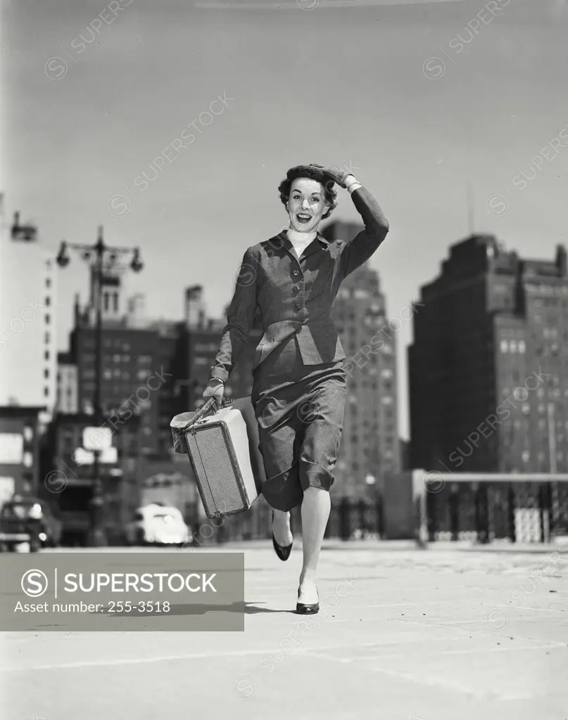 Vintage Photograph. Fashionable woman running on sidewalk carrying purse and small suitcase and holding hat on head