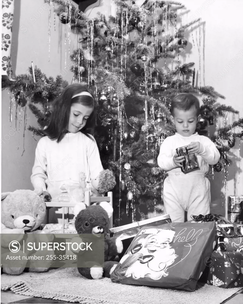 Girl playing with a doll and her brother opening a Christmas present beside her