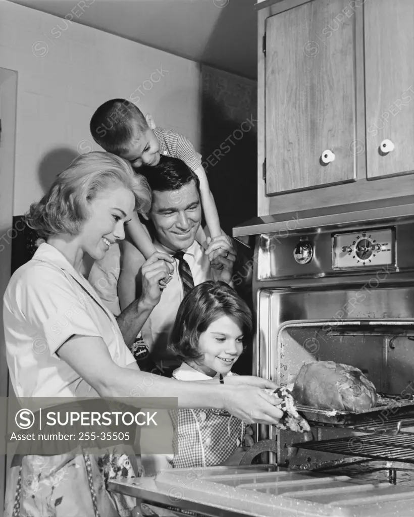Side profile of a mid adult woman putting a turkey into an oven with her family standing beside her