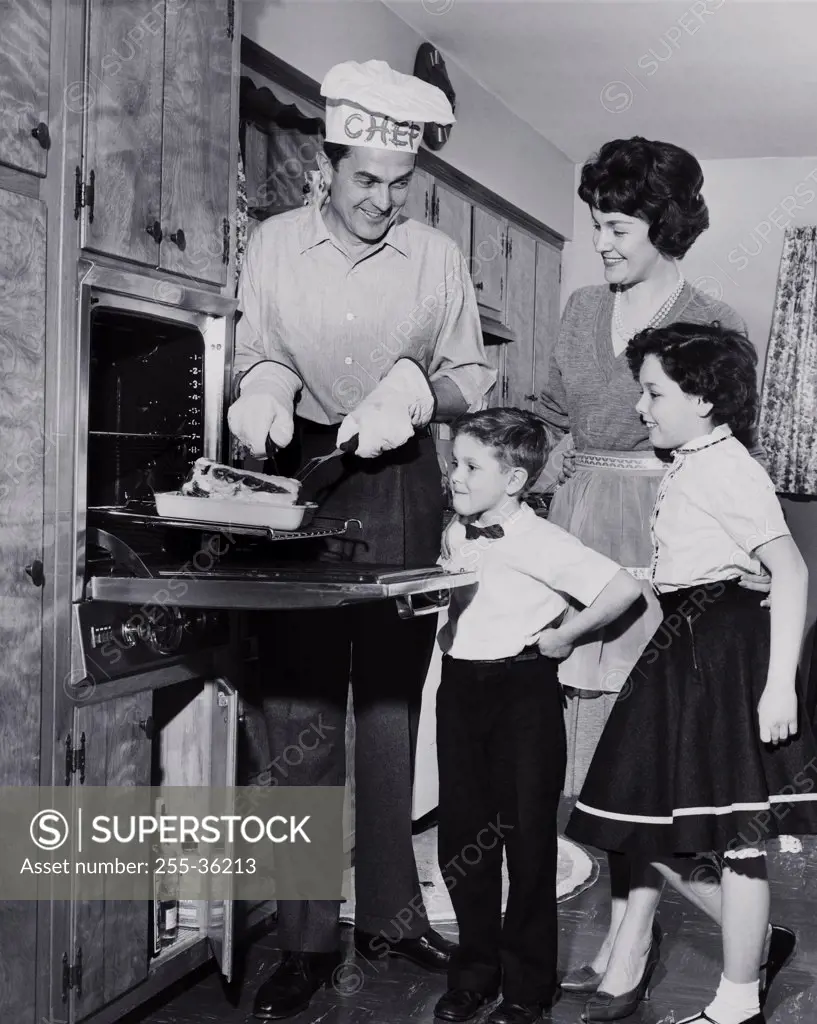 Mid adult man taking food out of an oven with his family standing beside him